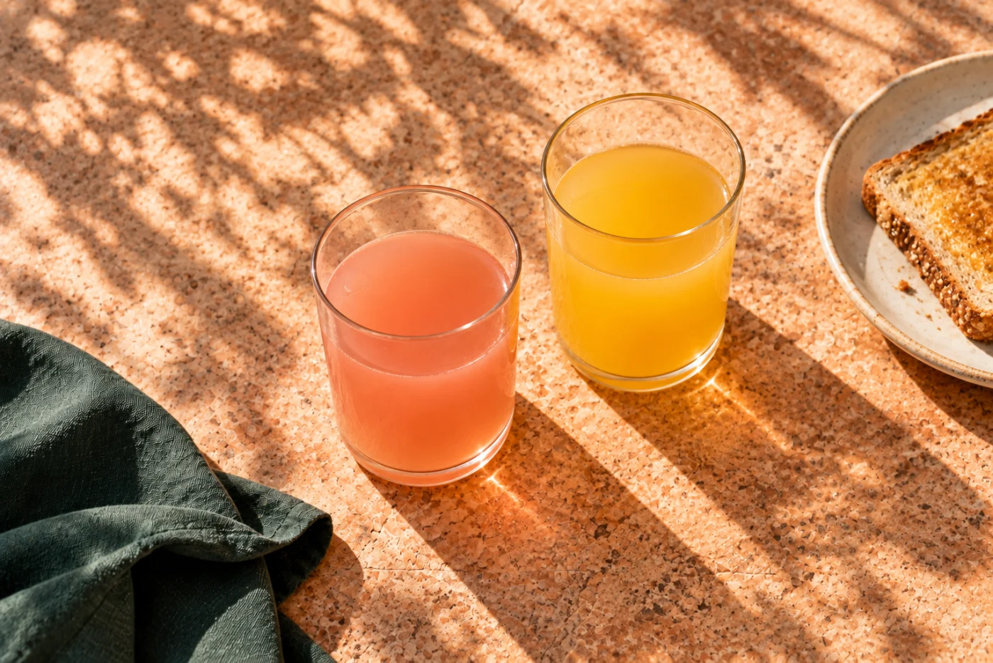 Pink and golden fruit-flavoured drinks without ice in ordinary glasses beside toast on a sunny table